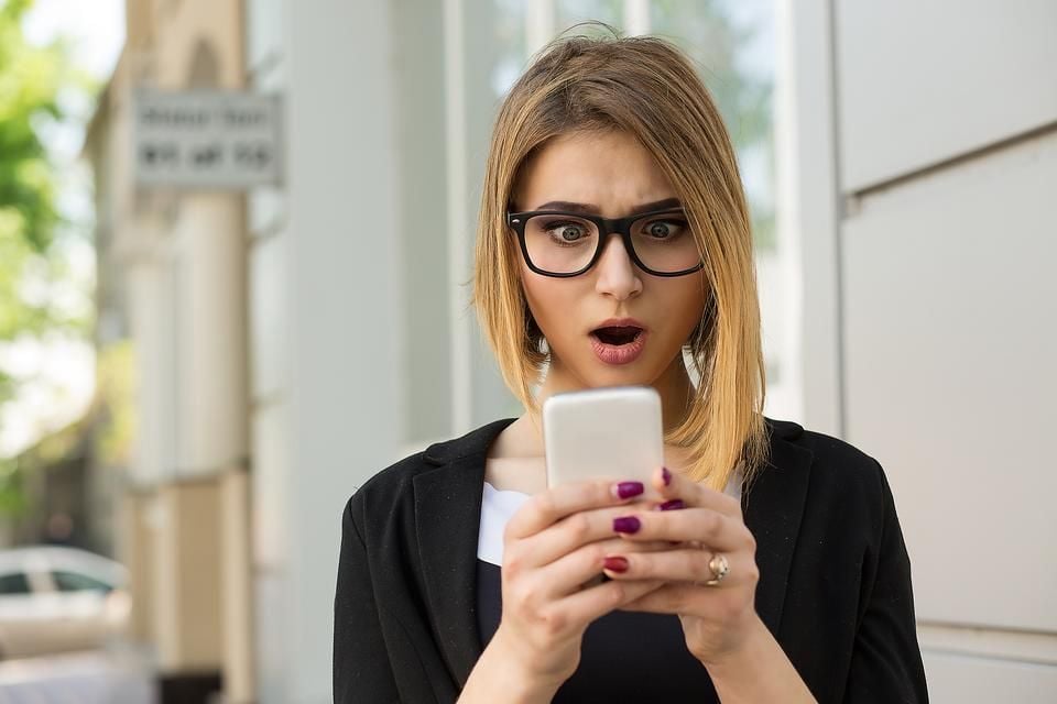 A woman with glasses and shoulder-length hair looks surprised while staring at her smartphone, standing outdoors near a building.