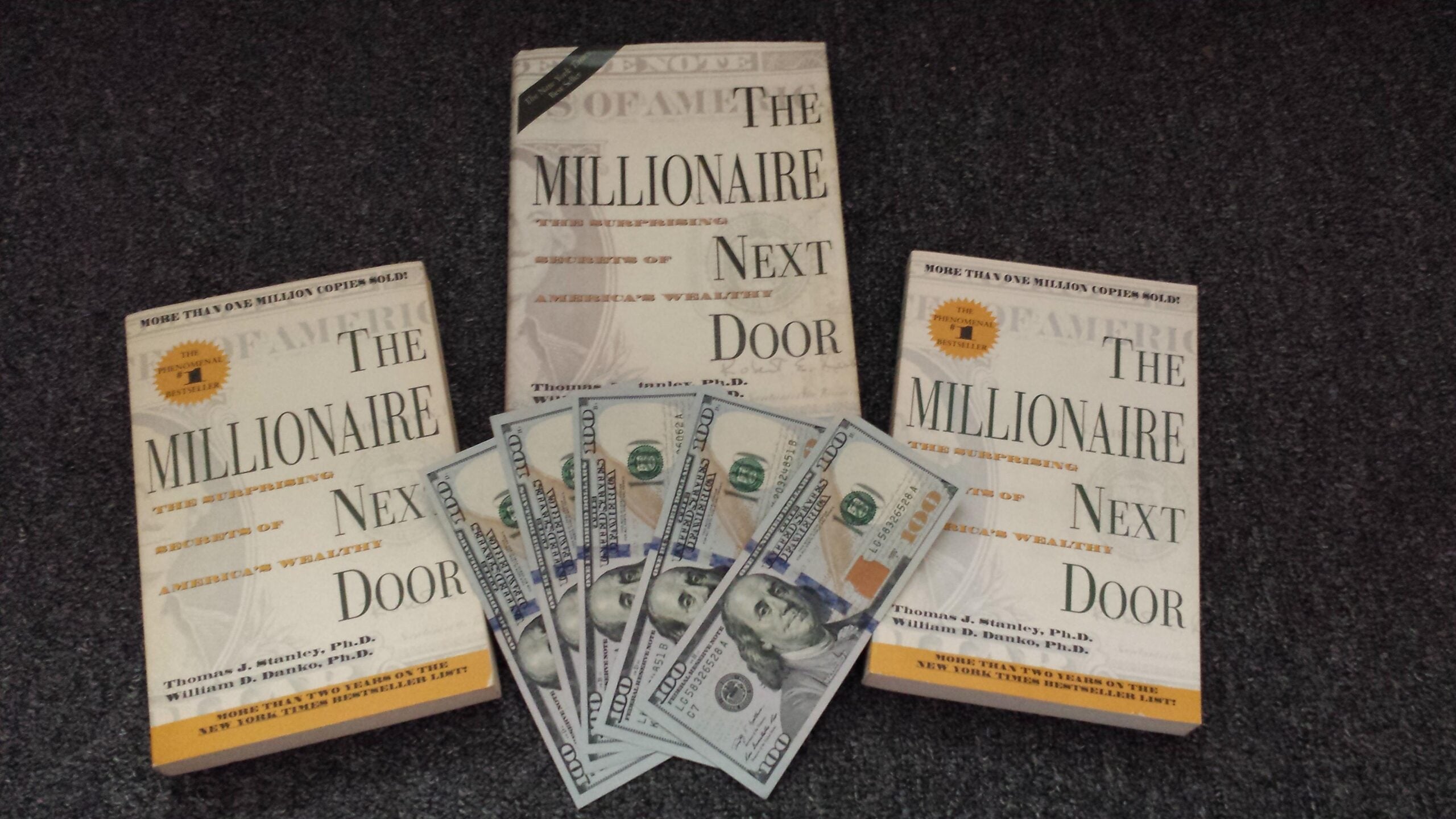Three copies of the book "The Millionaire Next Door" are displayed on a dark surface, with a fan of six 100-dollar bills arranged in front of the center book.