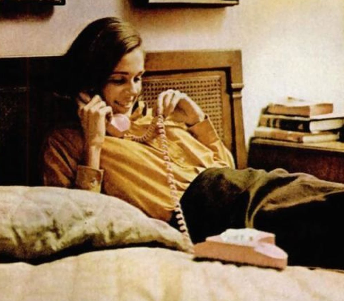 A woman reclines on a bed, smiling as she talks on a pink rotary telephone. There are books stacked on a bedside table behind her.