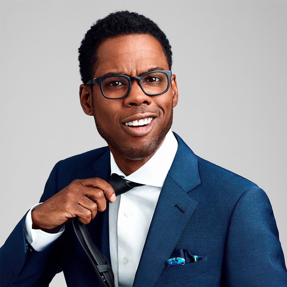 A man wearing glasses and a blue suit adjusts his black tie while smiling at the camera against a plain light gray background.