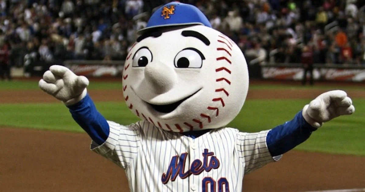 A person in a New York Mets mascot costume, featuring a large baseball head, striped jersey, and blue cap, smiles and waves on a baseball field with a blurred crowd in the background.