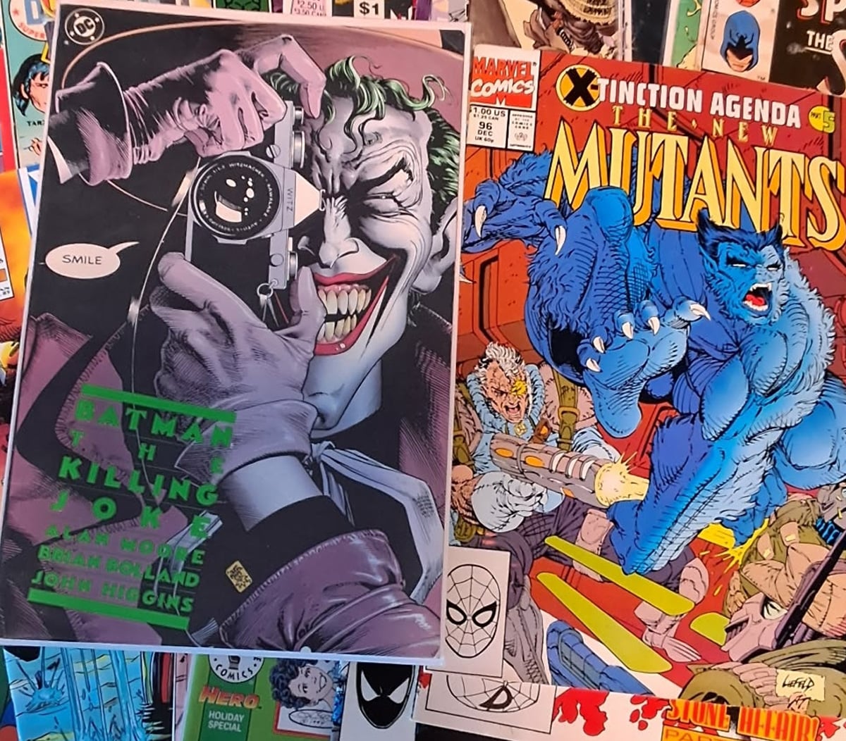 Two comic book covers: one of "Batman: The Killing Joke" featuring the Joker holding a camera and grinning, and another of "The New Mutants" with a blue beast attacking a group of heroes. Other comics are visible in the background.