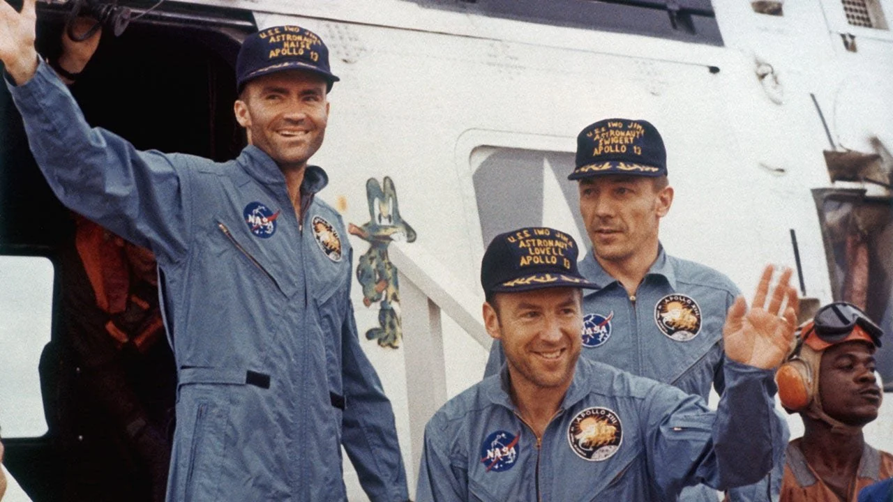 Three astronauts in blue NASA jumpsuits and navy caps wave and smile near a spacecraft hatch, with mission patches visible on their uniforms. A crew member in red gear is seen in the foreground.