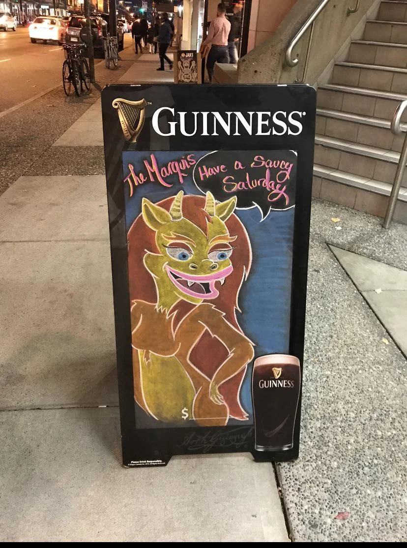 A sidewalk chalkboard sign features a colorful cartoon dragon holding a Guinness pint. The sign reads, "The Marquis Have a Saucy Saturday," with Guinness branding at the top and a pint illustration at the bottom.