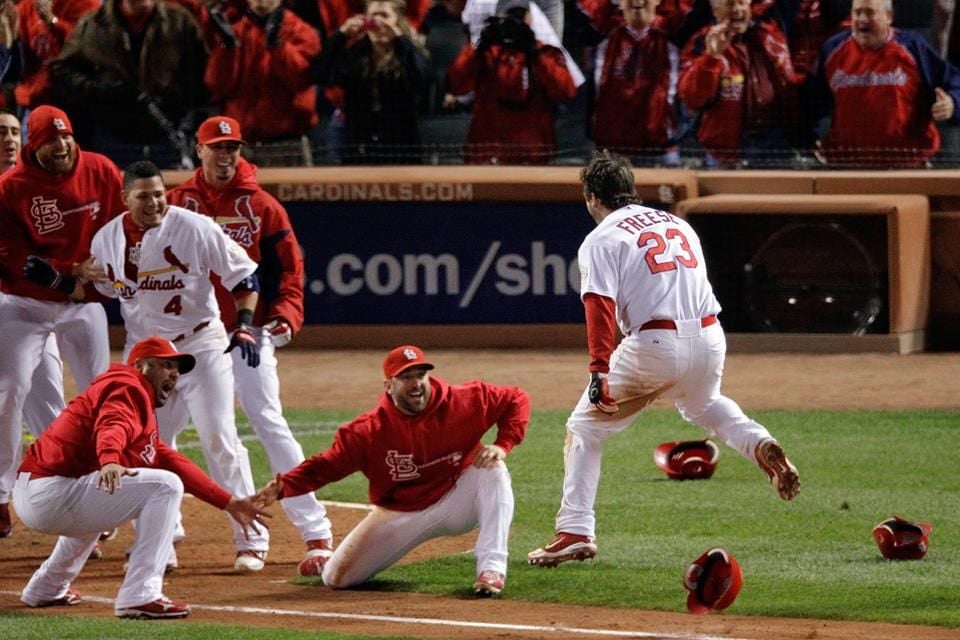 A baseball player wearing a "23" jersey celebrates at home plate as teammates in St. Louis Cardinals uniforms cheer and reach out to greet him, with hats scattered on the field.