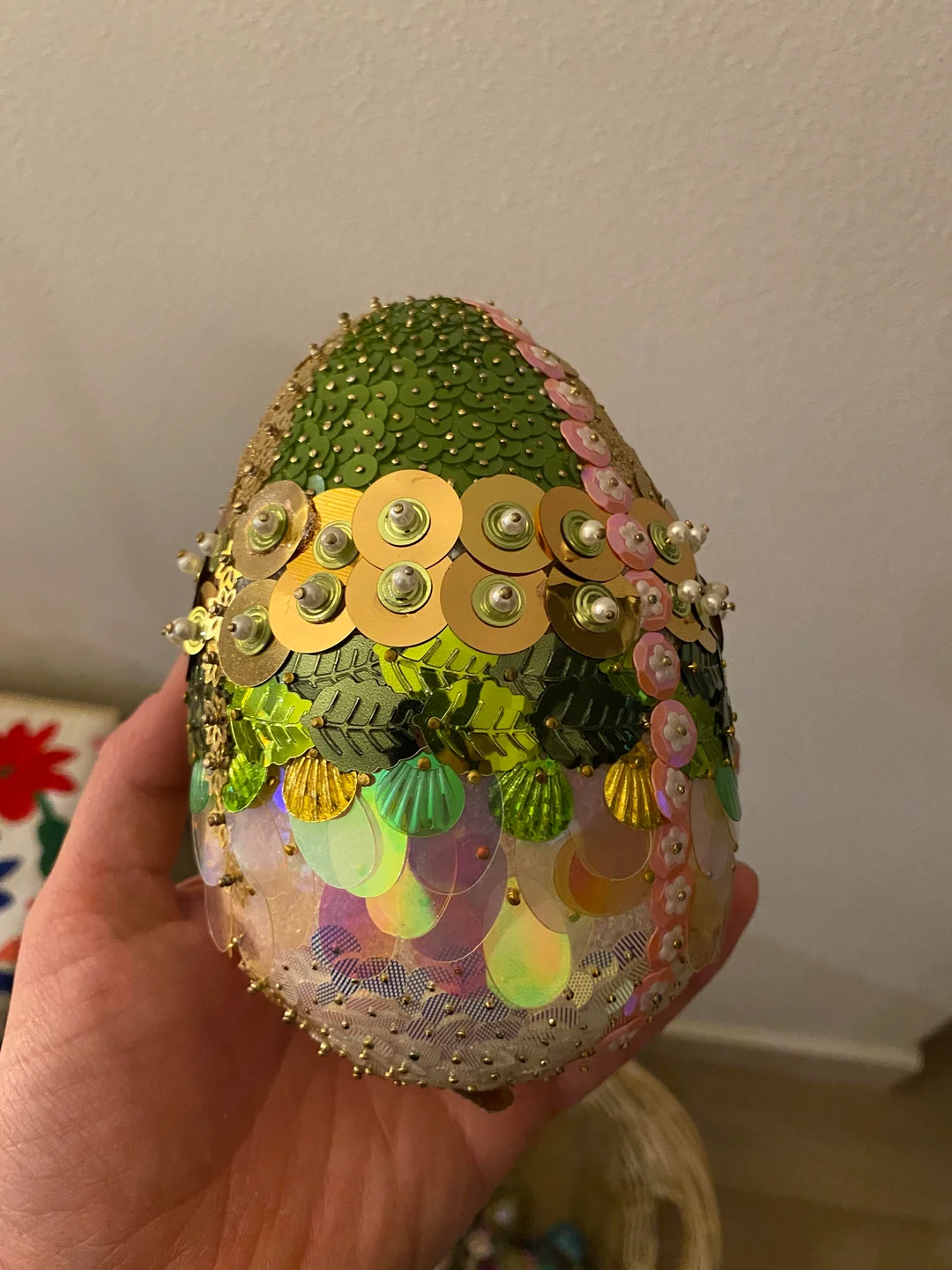 A hand holds a decorative egg covered in green, gold, pink, and iridescent sequins and beads, forming a shimmering textured pattern. The background is neutral with part of a patterned surface visible.