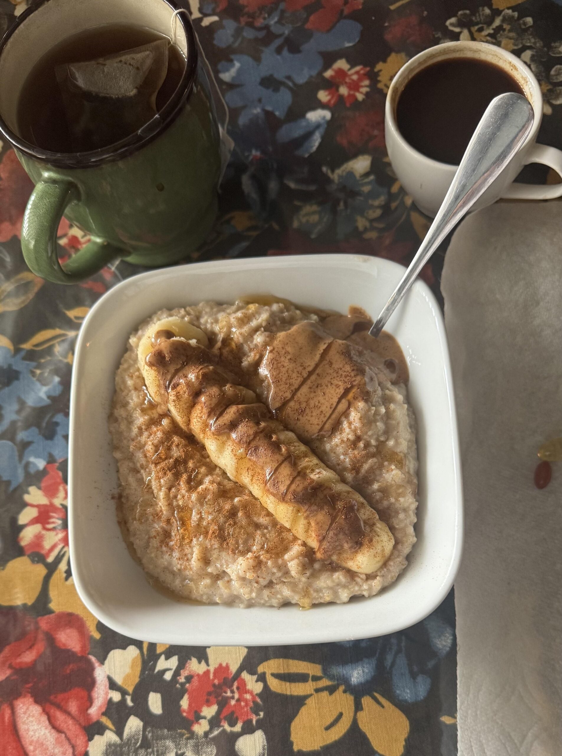 A bowl of oatmeal topped with a sliced banana, a dollop of nut butter, and cinnamon sits on a floral tablecloth. Nearby are a mug of tea with a teabag and a cup of coffee, with a napkin and vitamins on the side.
