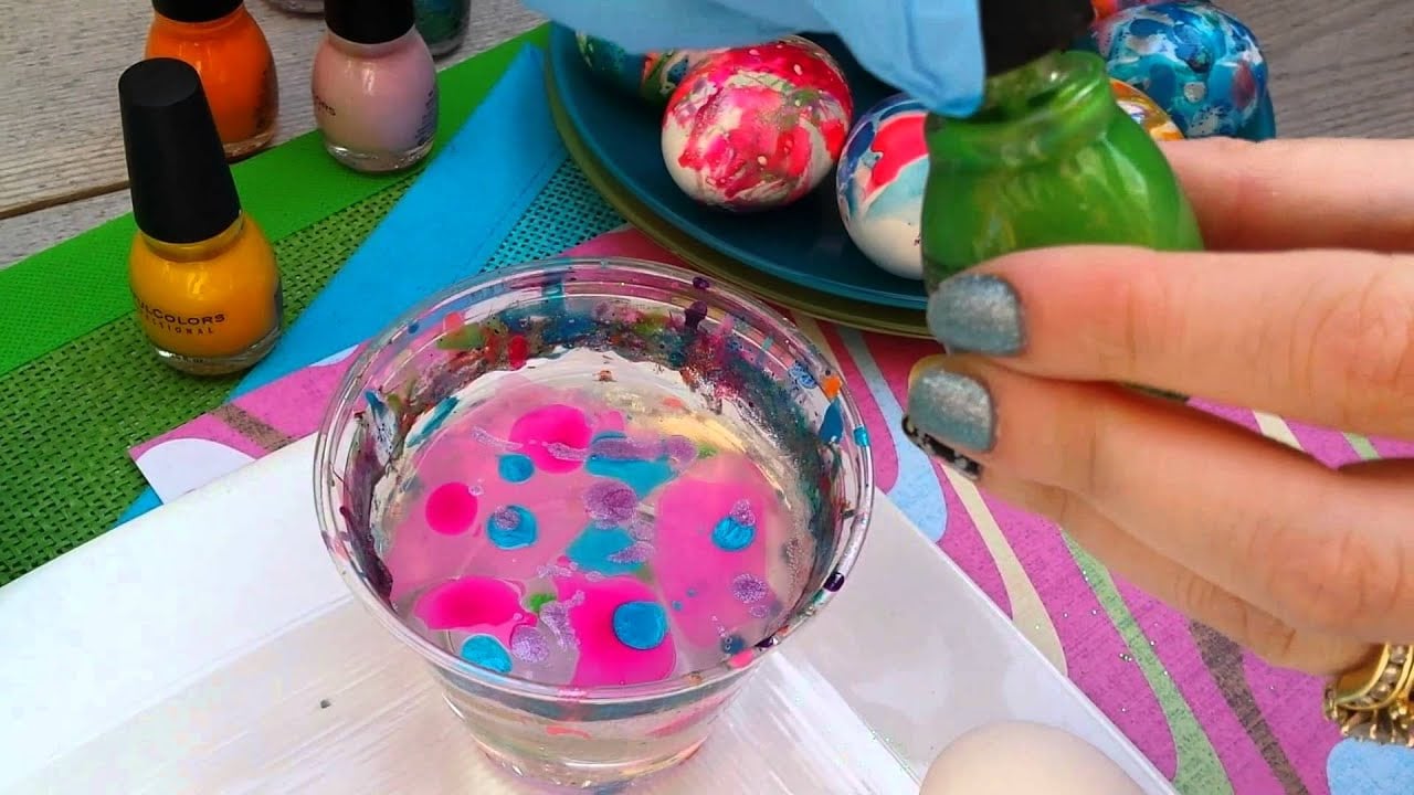 A hand with glittery blue nail polish drops green polish into a glass of water with floating pink and blue polish, while marbled Easter eggs and nail polish bottles are seen nearby.