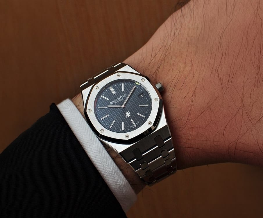 A close-up of a man's wrist wearing a luxury stainless steel wristwatch with a dark blue face and silver hour markers, paired with a black suit and white cuff.
