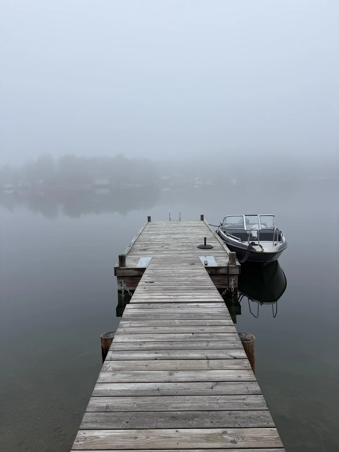 A wooden dock extends into a calm, foggy lake with a small motorboat moored on the right side. Trees and buildings are faintly visible in the misty background.