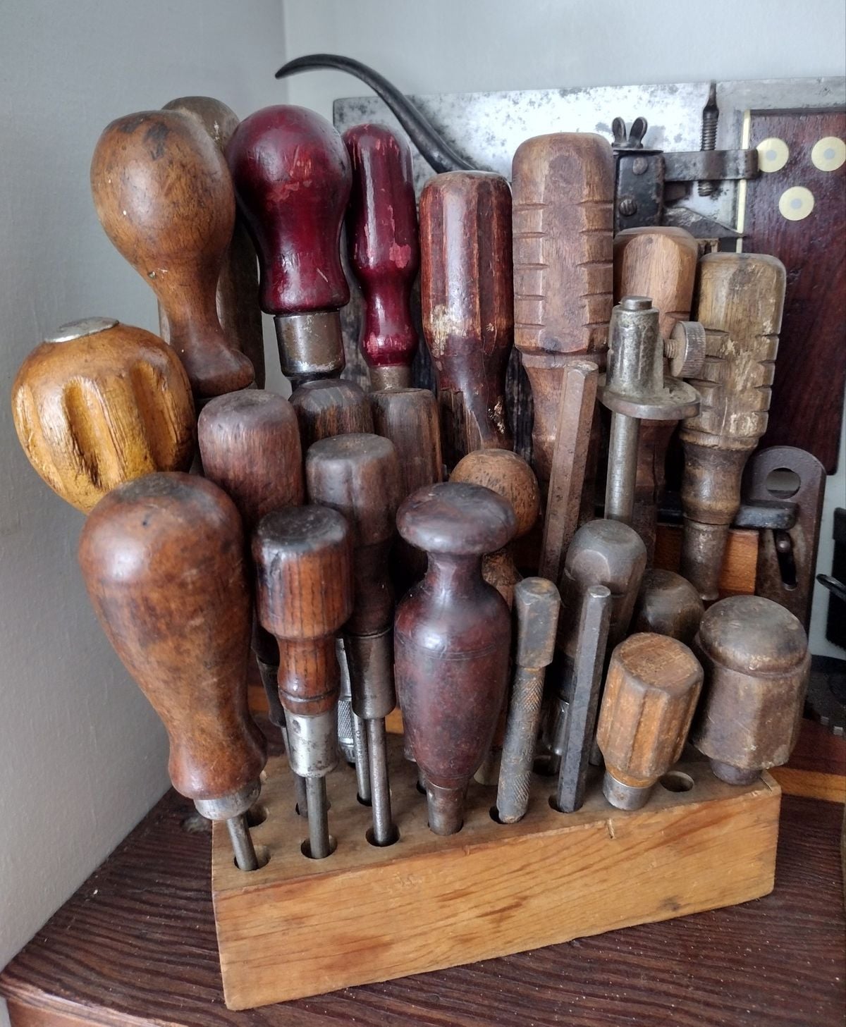 A wooden block holds a variety of vintage screwdrivers and hand tools with worn wooden handles, arranged upright in holes on a workbench.