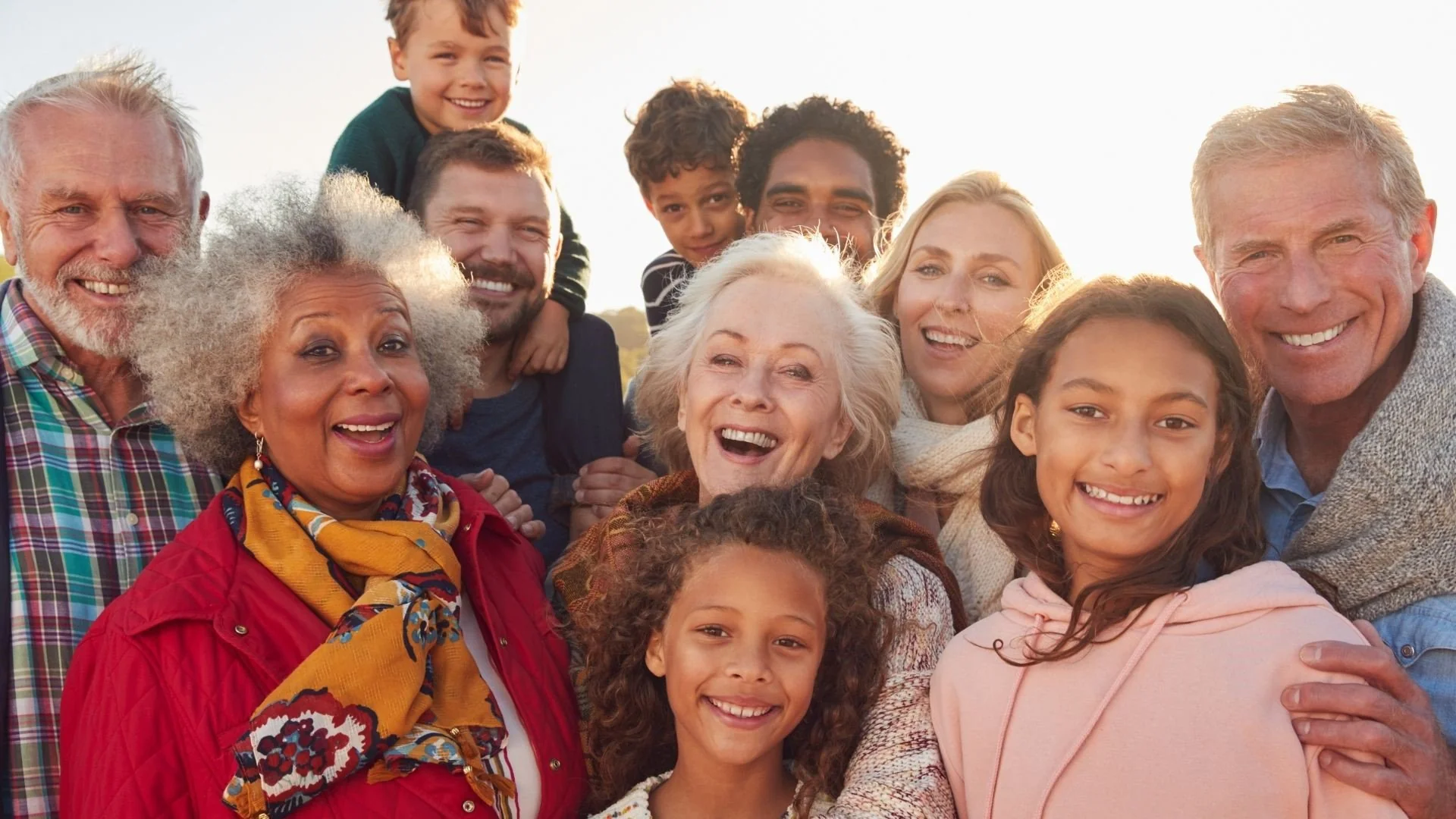 A diverse, multi-generational group of people smiling and posing closely together outdoors, with warm sunlight in the background. The group includes children, adults, and seniors, all appearing happy and cheerful.