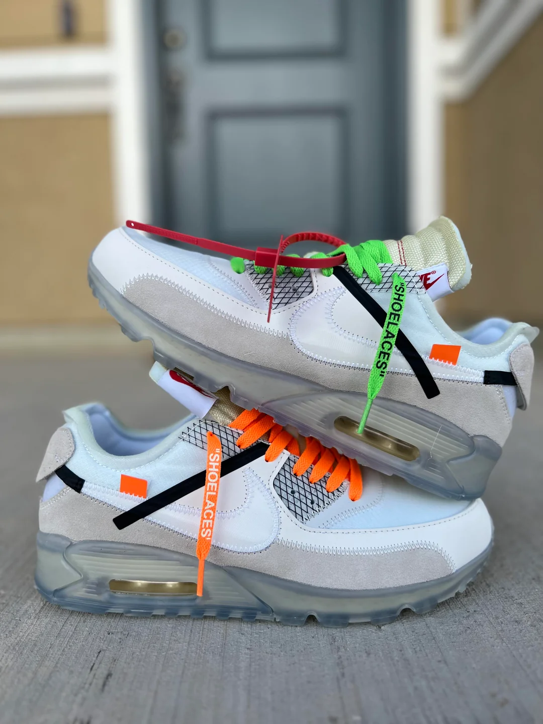 A pair of white and gray sneakers stacked sideways, featuring translucent soles and colorful shoelaces—one pair orange, the other green—each labeled with "SHOELACES" text. The background shows a blurred door and wall.