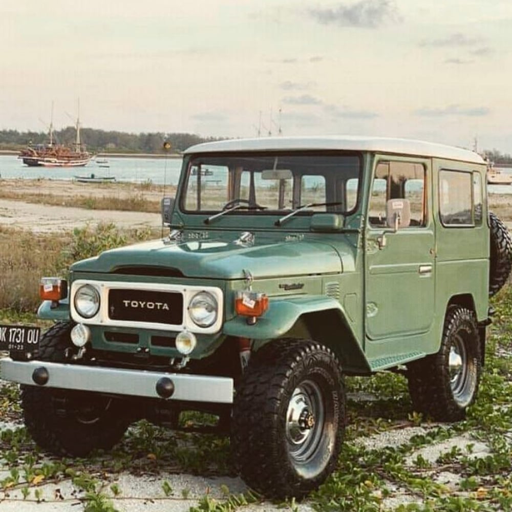 A vintage green Toyota Land Cruiser is parked on grassy terrain near a sandy beach, with several boats visible in the water and a cloudy sky in the background.