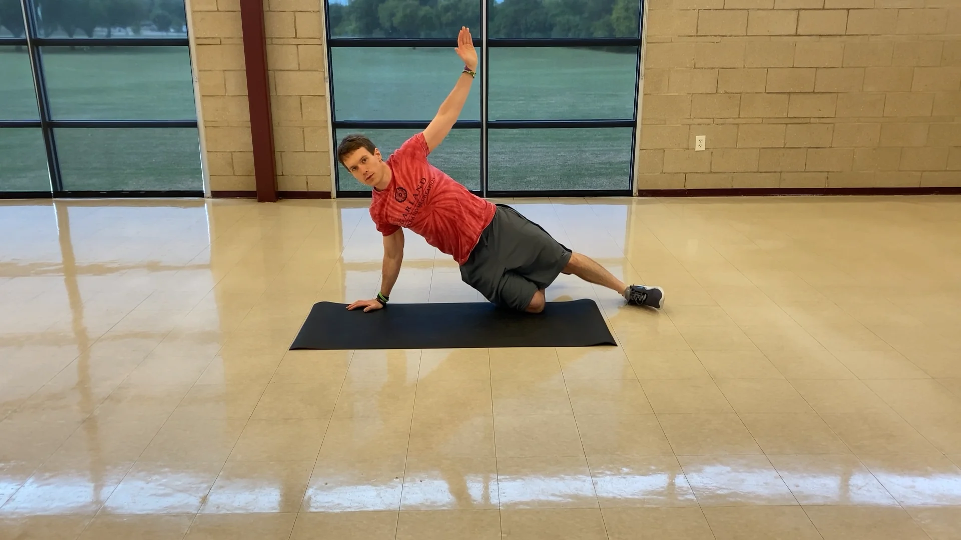 A man in a red shirt and gray shorts performs a side plank on a black mat indoors, with one knee bent on the floor and the other leg extended, raising one arm upward. Large windows and a brick wall are in the background.
