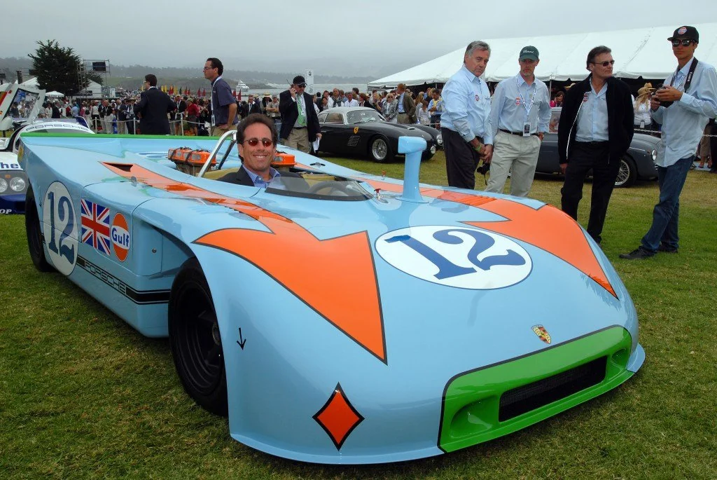 A smiling man sits in a vintage blue and orange race car with the number 12, surrounded by people at an outdoor car event with tents and classic cars in the background.