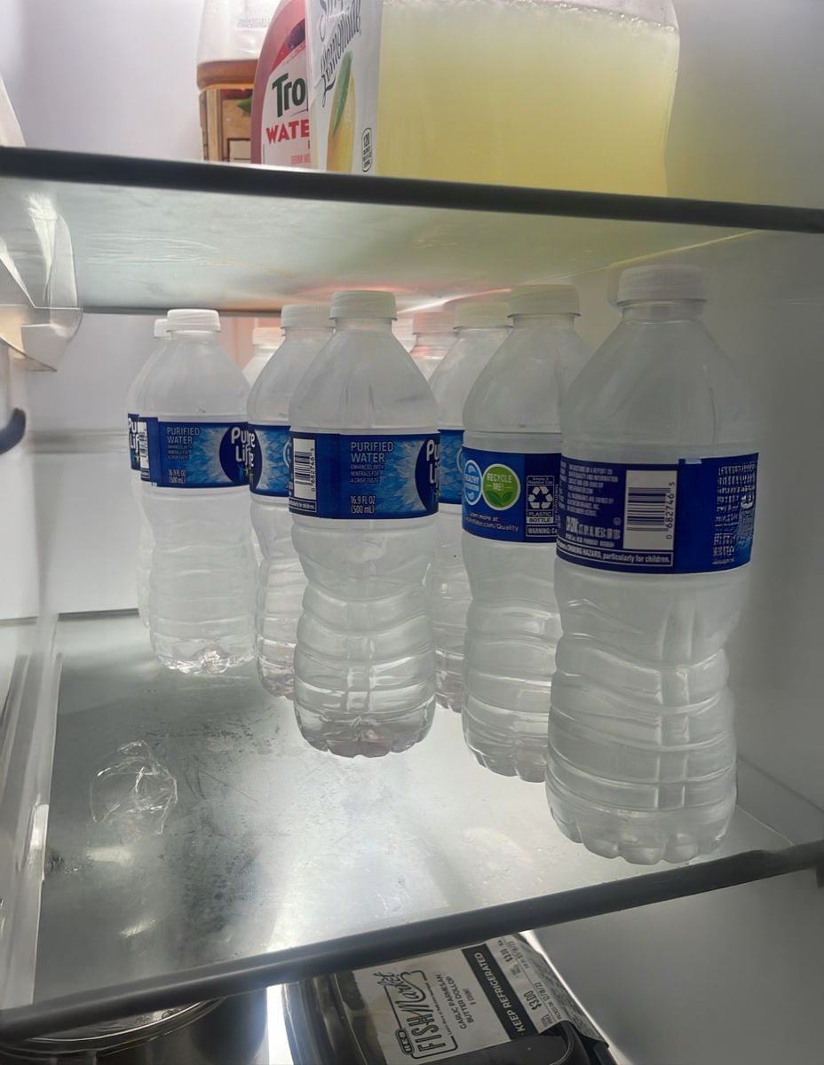 Several partially filled plastic water bottles sit on a refrigerator shelf, with a large container of yellow liquid and other items above, and a metal tray with a black lid below. The fridge is brightly lit.