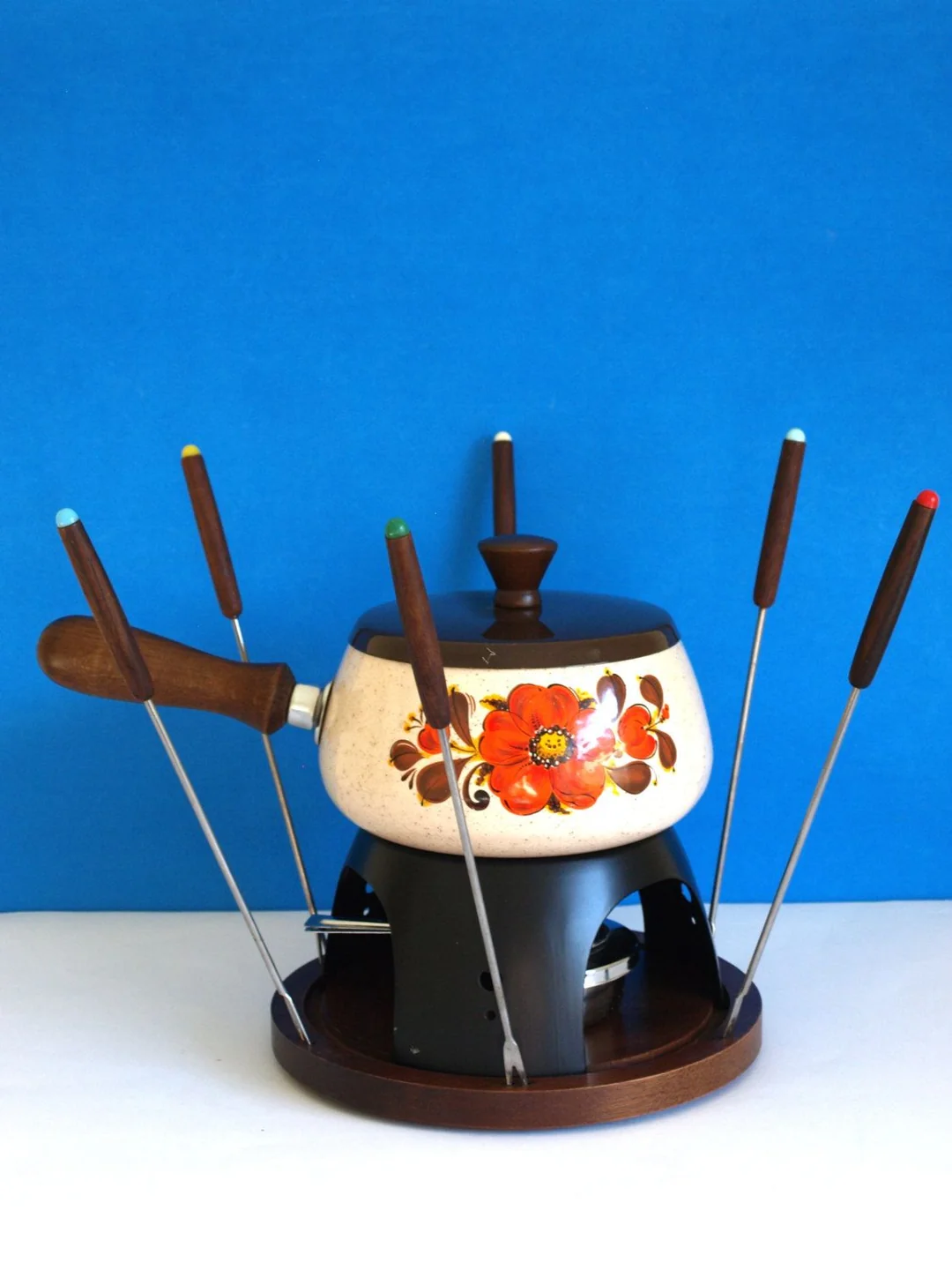A vintage fondue pot with a painted red and orange flower design, wooden handle and lid, and six fondue forks with colored tips, arranged in a circular stand against a blue background.