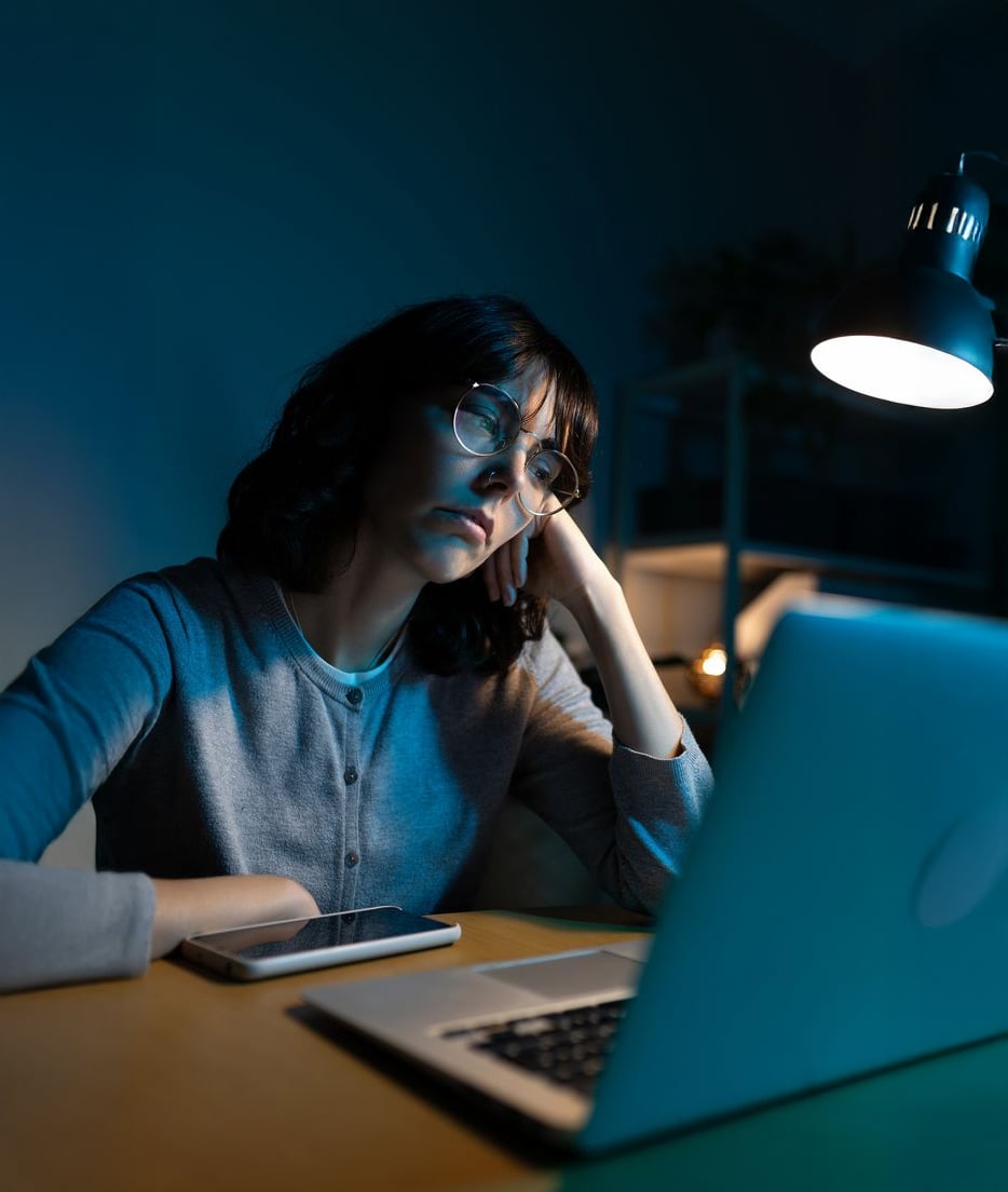 A woman wearing glasses sits at a desk, resting her head on her hand and looking tiredly at a laptop screen, with a phone and a lamp nearby in a dimly lit room.