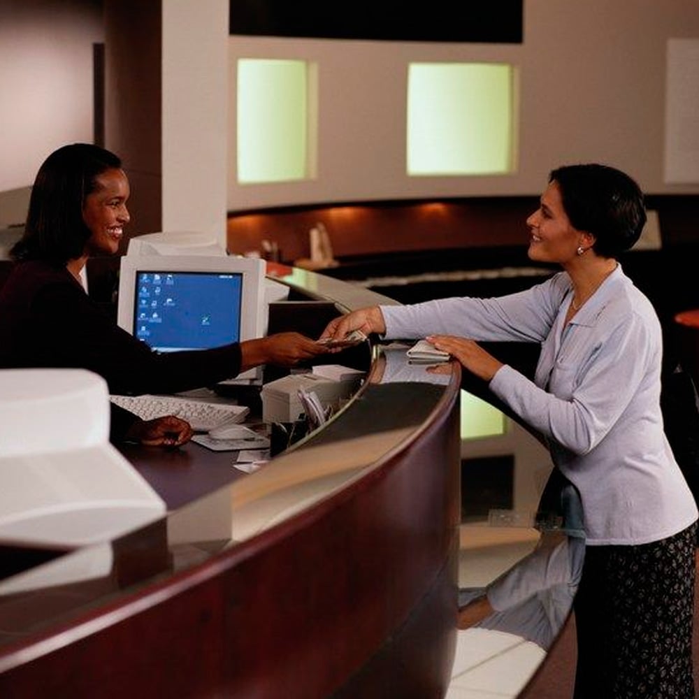 A receptionist behind a desk hands documents to a smiling woman standing on the other side. Both women appear friendly and are interacting in a modern, well-lit office or hotel lobby.