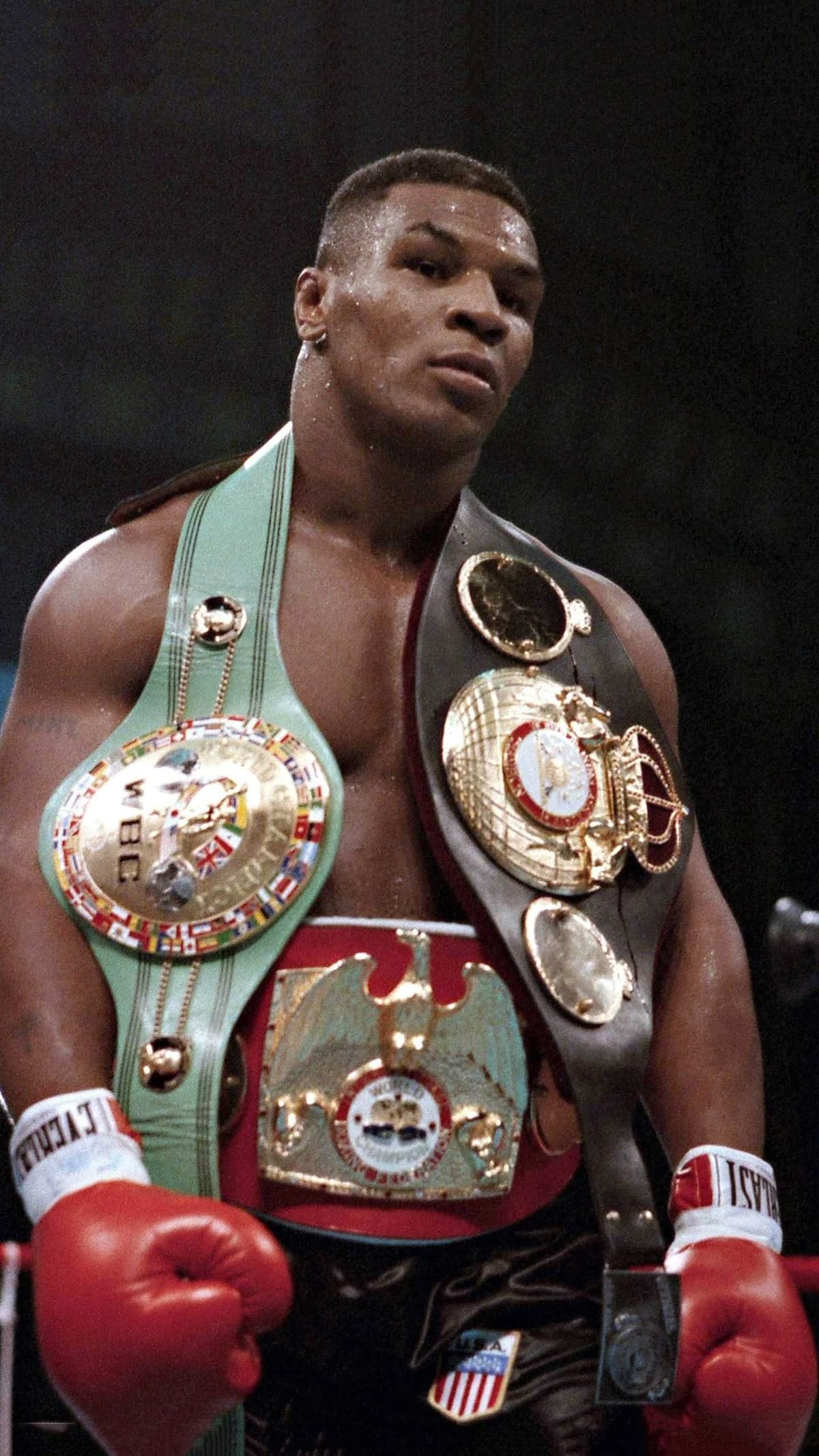 A male boxer wearing red gloves stands in the ring with three championship belts draped over his shoulders and waist, looking serious after a match.