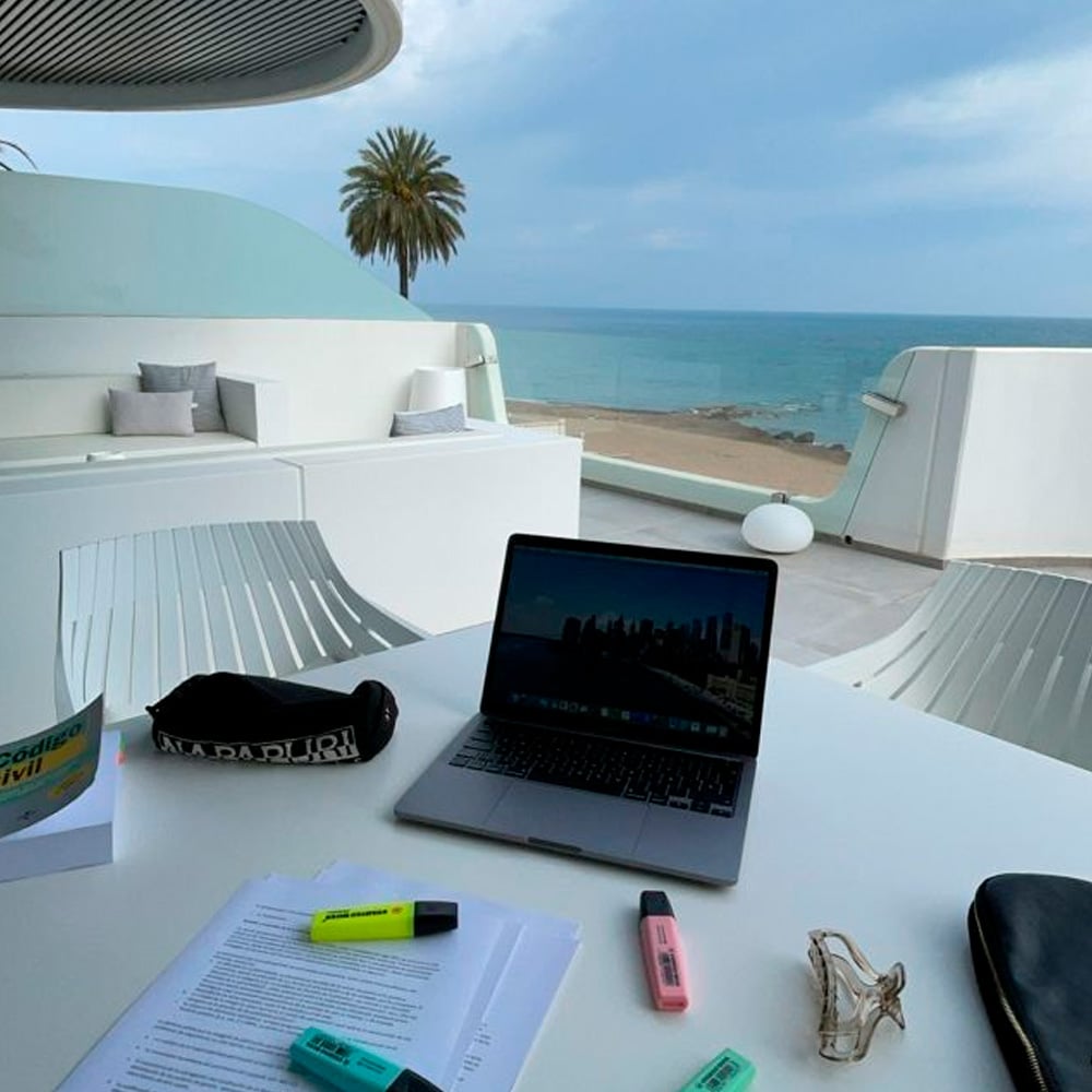 A laptop, papers, highlighters, glasses, and a pencil case sit on a white table on a terrace overlooking a sandy beach and blue ocean, with modern white furniture and a palm tree in the background.