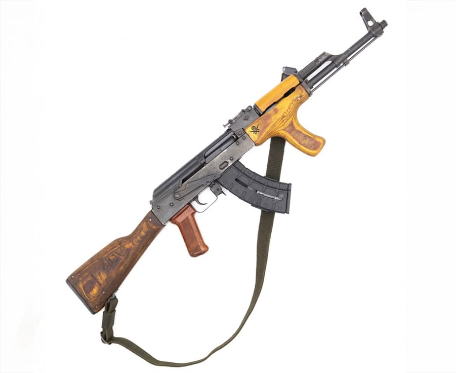 A Kalashnikov-style assault rifle with a wooden stock, wooden handguard, and pistol grip, featuring a black metal receiver, a curved magazine, and an attached green shoulder sling.
