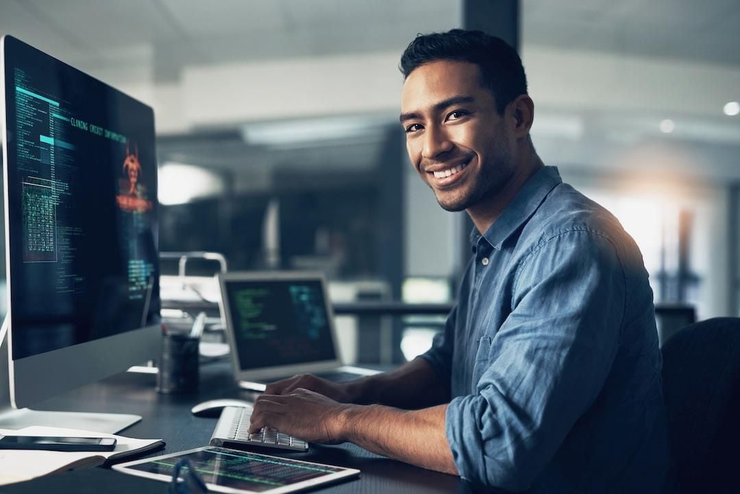 A man in a blue shirt smiles while working at a computer with code on the screen in a modern office setting. A tablet and other equipment are on the desk in front of him.