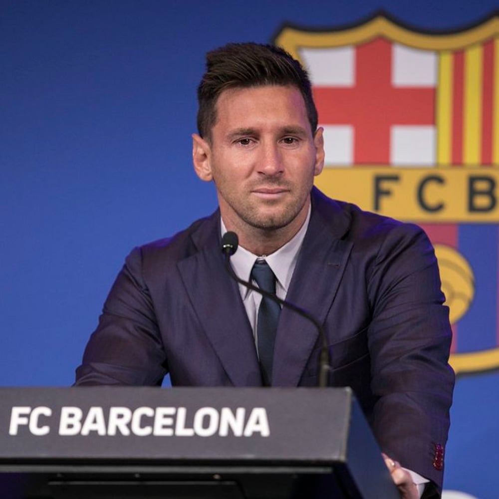 A man in a dark suit and tie sits at a podium labeled "FC BARCELONA" with a serious expression, in front of a blue background featuring part of the FC Barcelona logo.