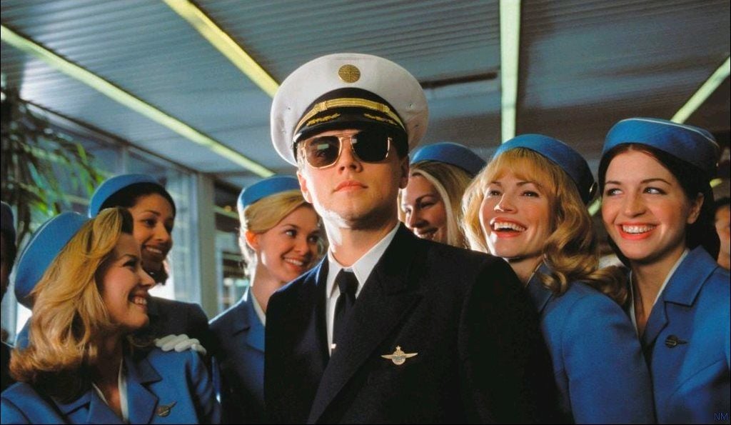 A man in a pilot uniform and sunglasses stands confidently, surrounded by smiling flight attendants in blue uniforms and matching hats. The group appears to be in an airport setting.