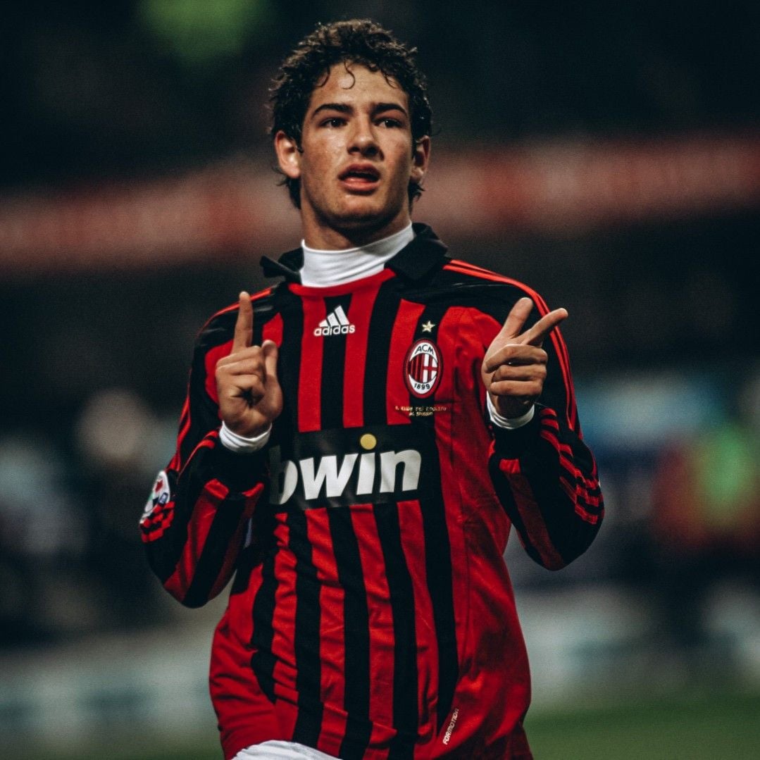 A soccer player in an AC Milan jersey gestures with both hands raised and index fingers pointing up during a match. He has curly hair and is wearing a red and black striped shirt with the "bwin" sponsor.