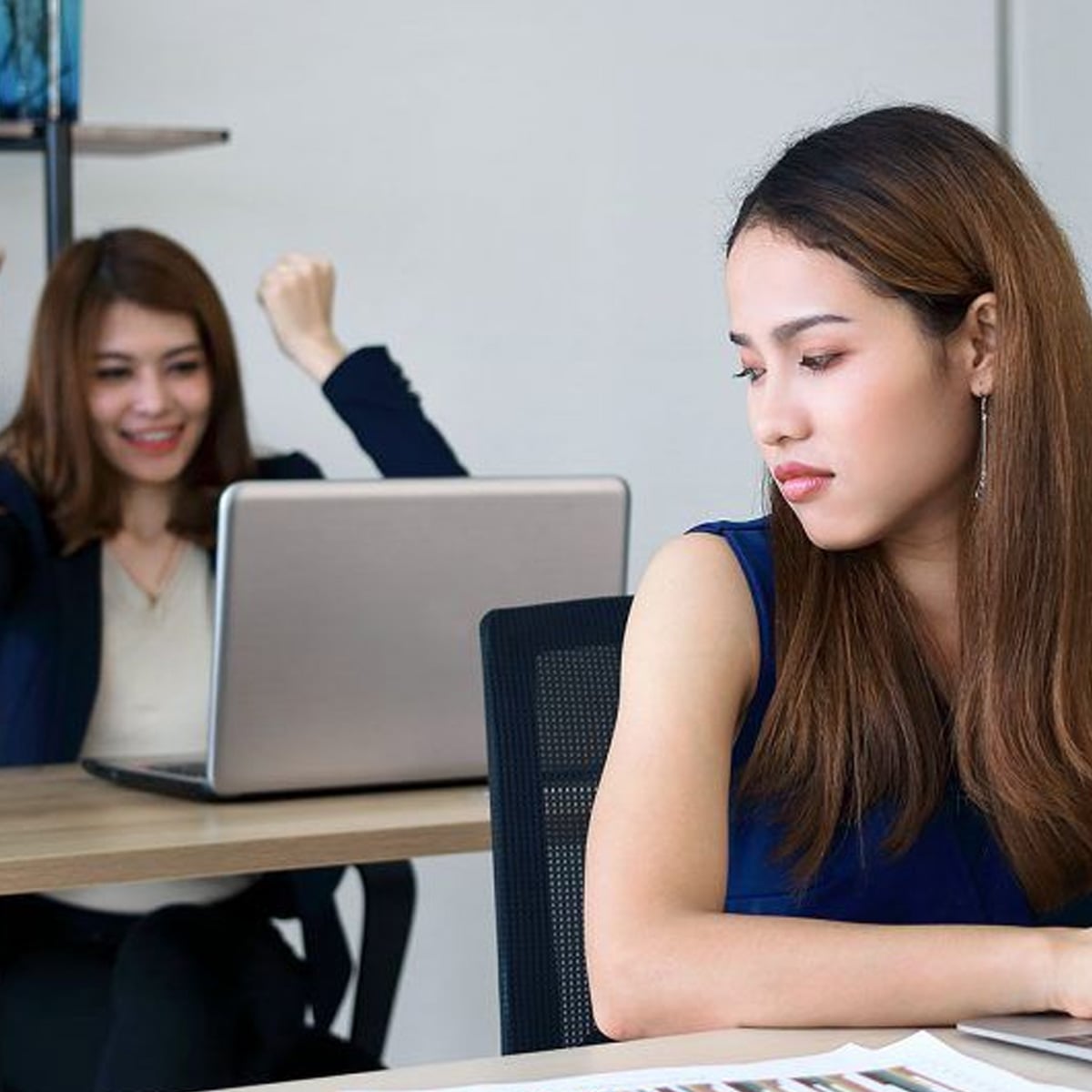 Two women are in an office. One woman in the foreground looks upset or disappointed, while the woman in the background is smiling and raising her arms in celebration in front of her laptop.