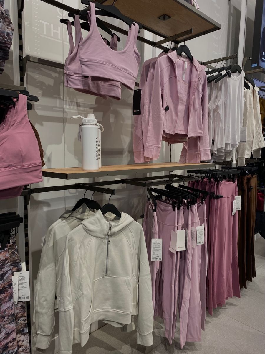 A clothing store display with pink and white activewear, including sports bras, zip-up jackets, sweatshirts, and pants neatly hung on black hangers, with a white water bottle on the shelf.