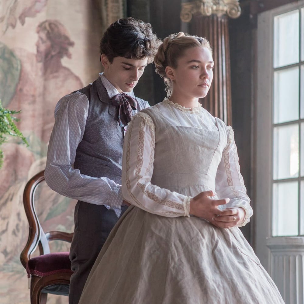 Two young people in period clothing stand indoors. The woman, dressed in a light, long-sleeved gown, looks ahead thoughtfully as the man behind her gently holds her hands. A vintage chair and tapestry are in the background.