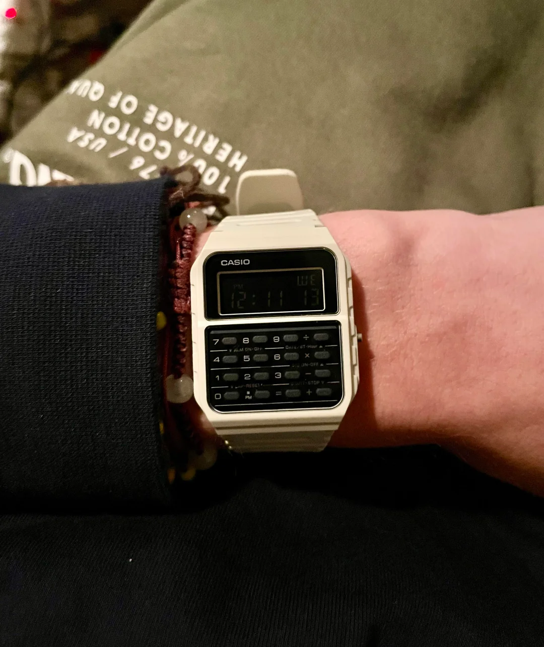 A person wears a white Casio calculator watch on their wrist. The time displayed is 12:11. The person has a dark sleeve and a bracelet, and green fabric with white text is visible in the background.
