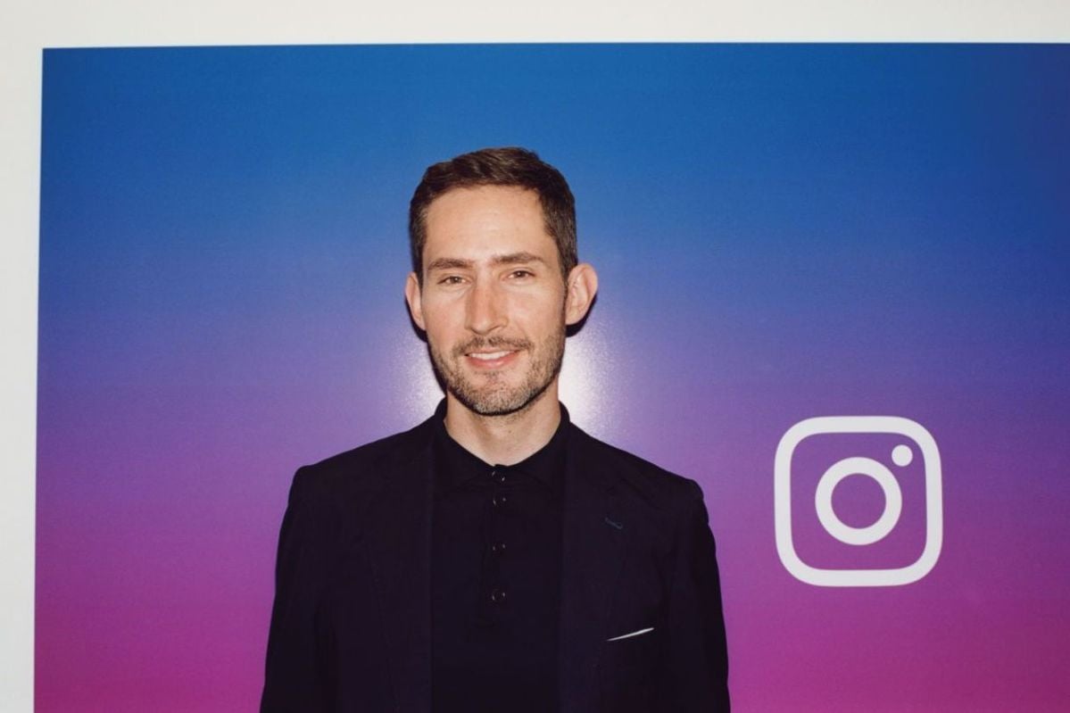 A man in a dark suit stands in front of a purple and blue gradient wall featuring the white Instagram logo. He is smiling slightly and looking at the camera.