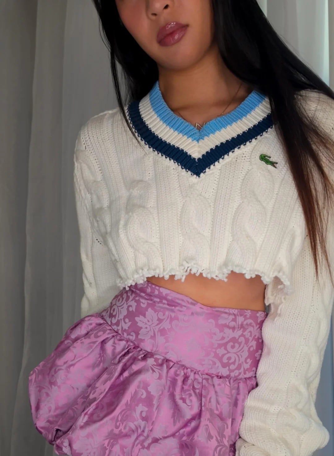 A woman poses indoors wearing a cropped white cable-knit sweater with a blue-trimmed V-neck and a light purple, floral-patterned skirt with ruffles. Her face is partially out of frame.