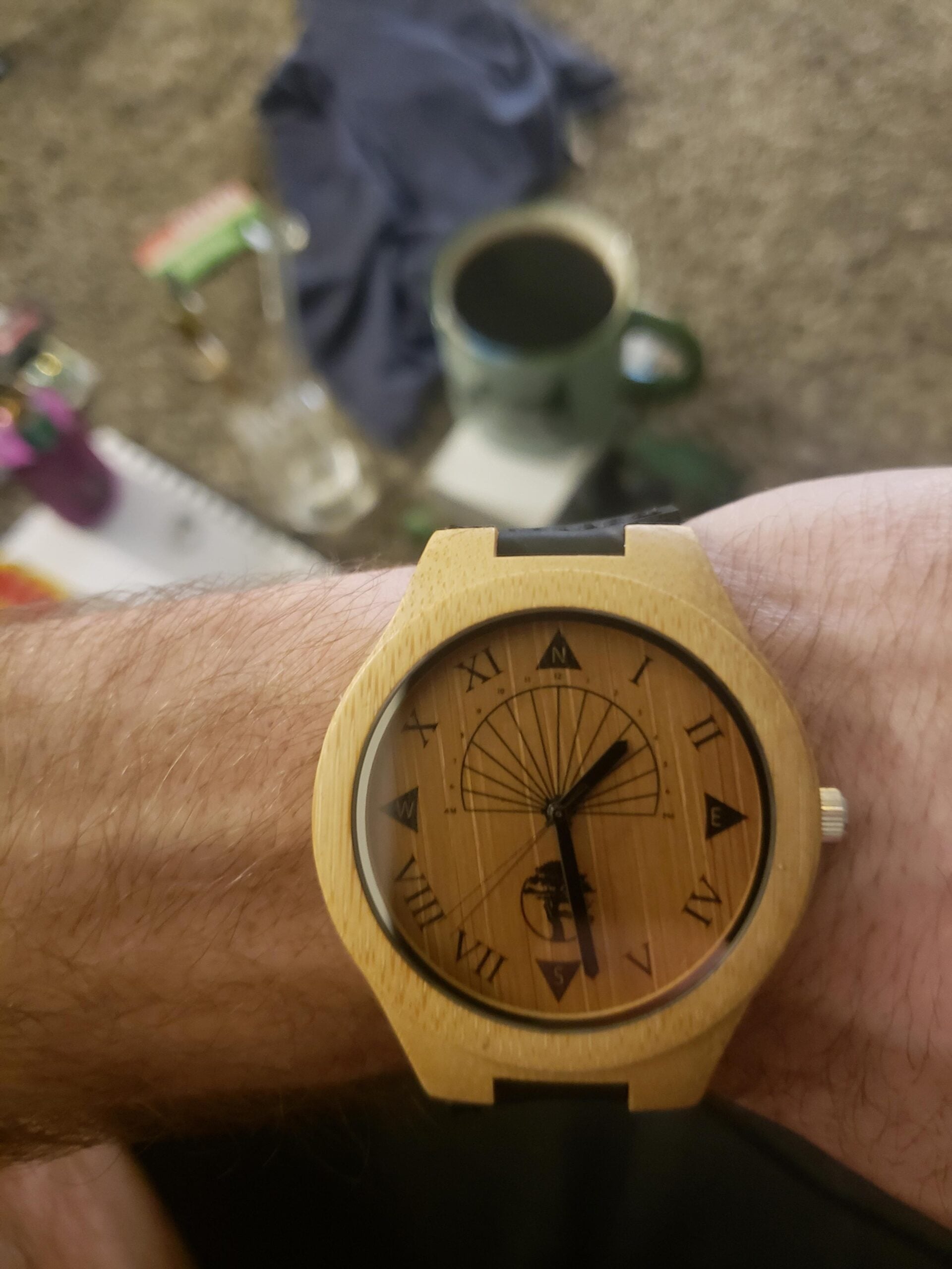 A person wearing a wooden watch with Roman numerals and an intricate design on the face. A mug, a cloth, and various items rest on a table in the blurred background.