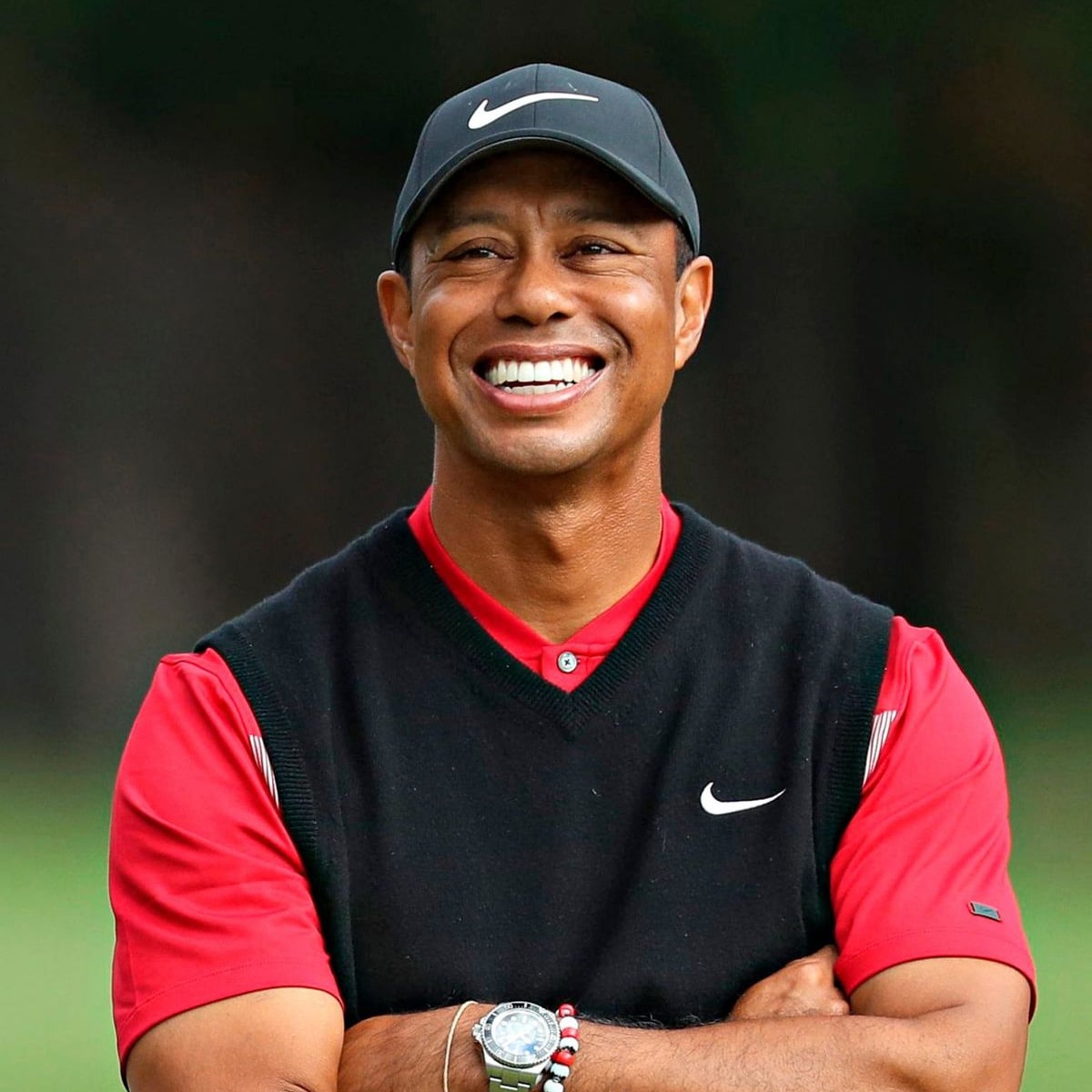 A man wearing a black Nike cap, black Nike vest over a red shirt, and a watch, smiling broadly with arms crossed, standing outdoors with a blurred background.