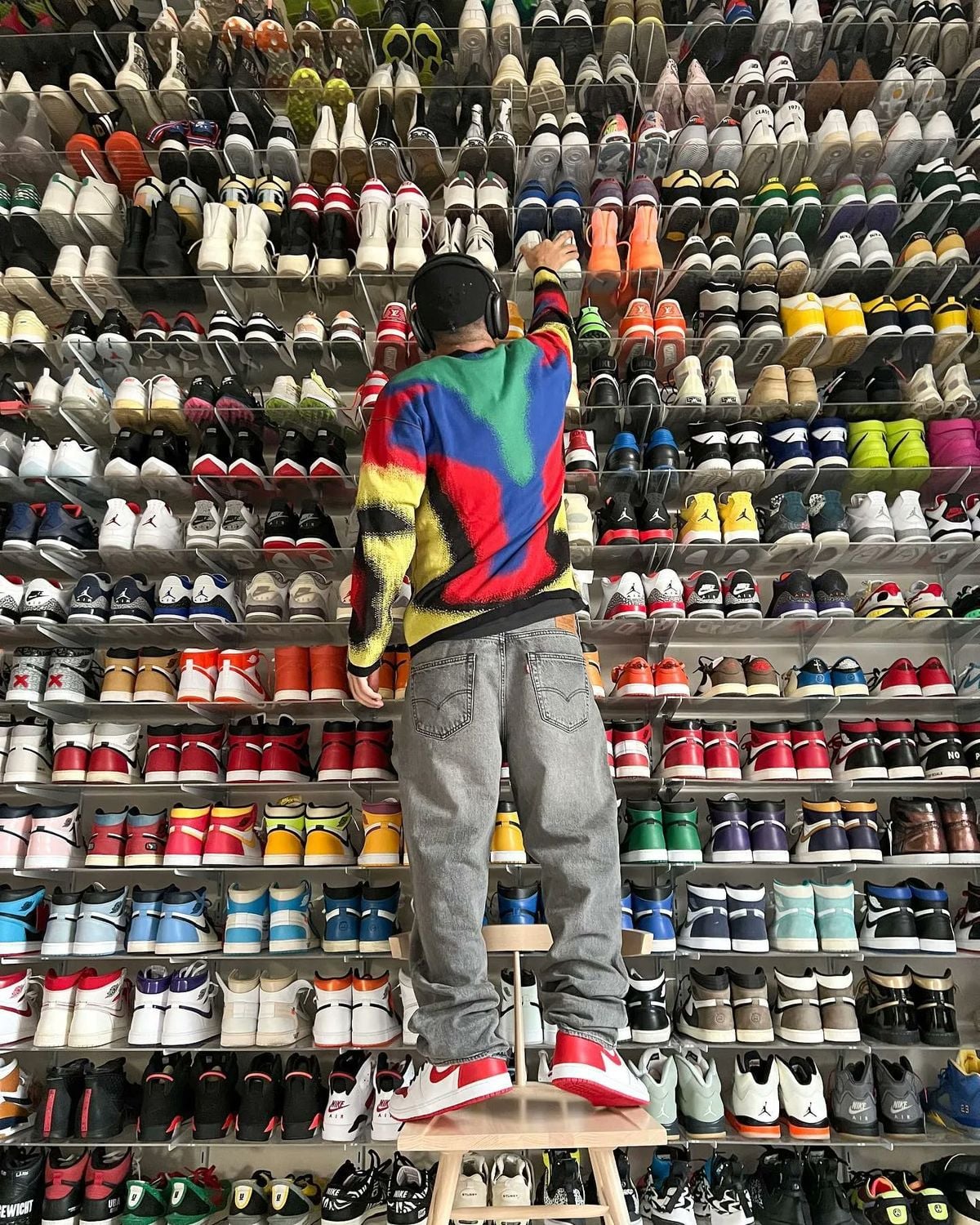 A person in a colorful sweater and jeans stands on a stool, reaching up to select a pair of sneakers from a large, floor-to-ceiling wall display filled with various colorful sneakers.