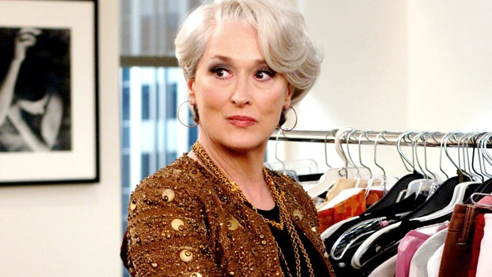 A woman with short gray hair and hoop earrings, wearing a gold embellished jacket, stands in front of a clothing rack, looking to the side in a stylish, modern office setting.