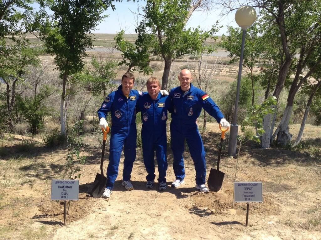 Three astronauts in blue space suits stand outdoors on either side of a patch of dirt, each holding a shovel, with small trees and name signs behind them, against a background of trees and open landscape.