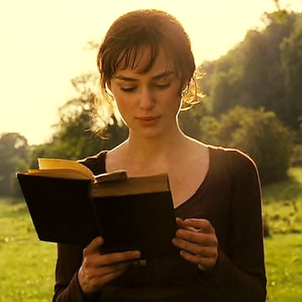 A woman with brown hair reads a book outdoors in a sunlit field, with greenery and trees in the background. She wears a simple brown dress and appears focused on the book.