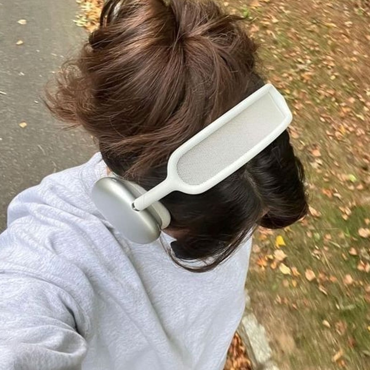 Person wearing large over-ear headphones and a light-colored sweatshirt, walking outdoors on a path lined with grass and fallen leaves, viewed from above.