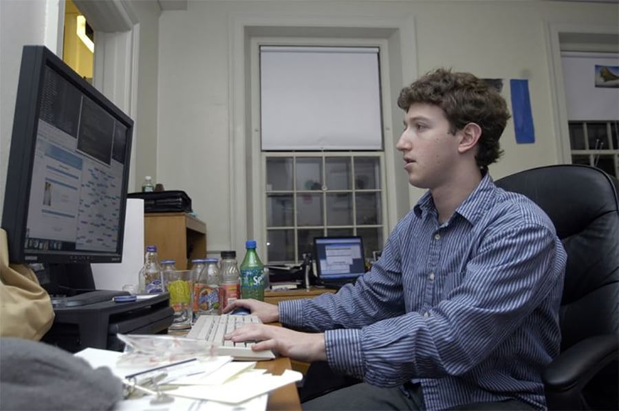 A young man in a striped shirt sits at a desk using a computer with two monitors. The desk is cluttered with bottles, papers, and office supplies, and a window is visible in the background.