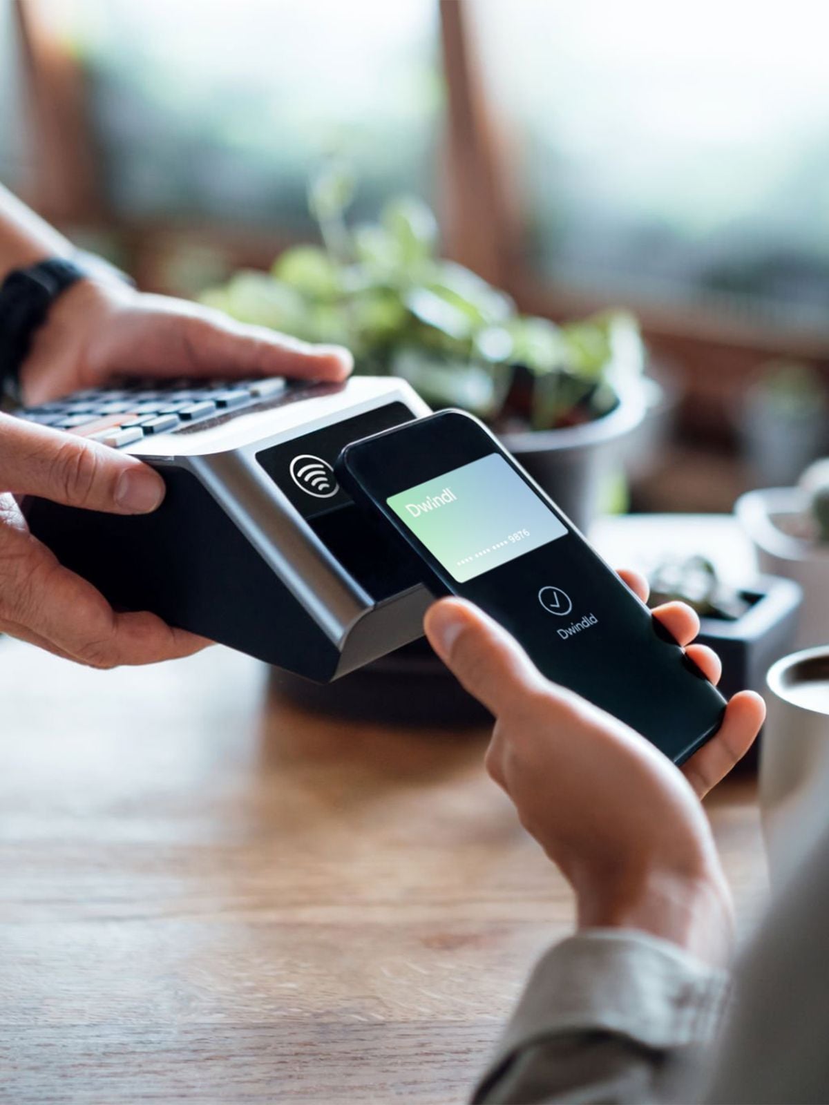 A person holds a smartphone near a wireless card reader to make a contactless payment, while another person holds the device; the background is softly blurred with plants visible.