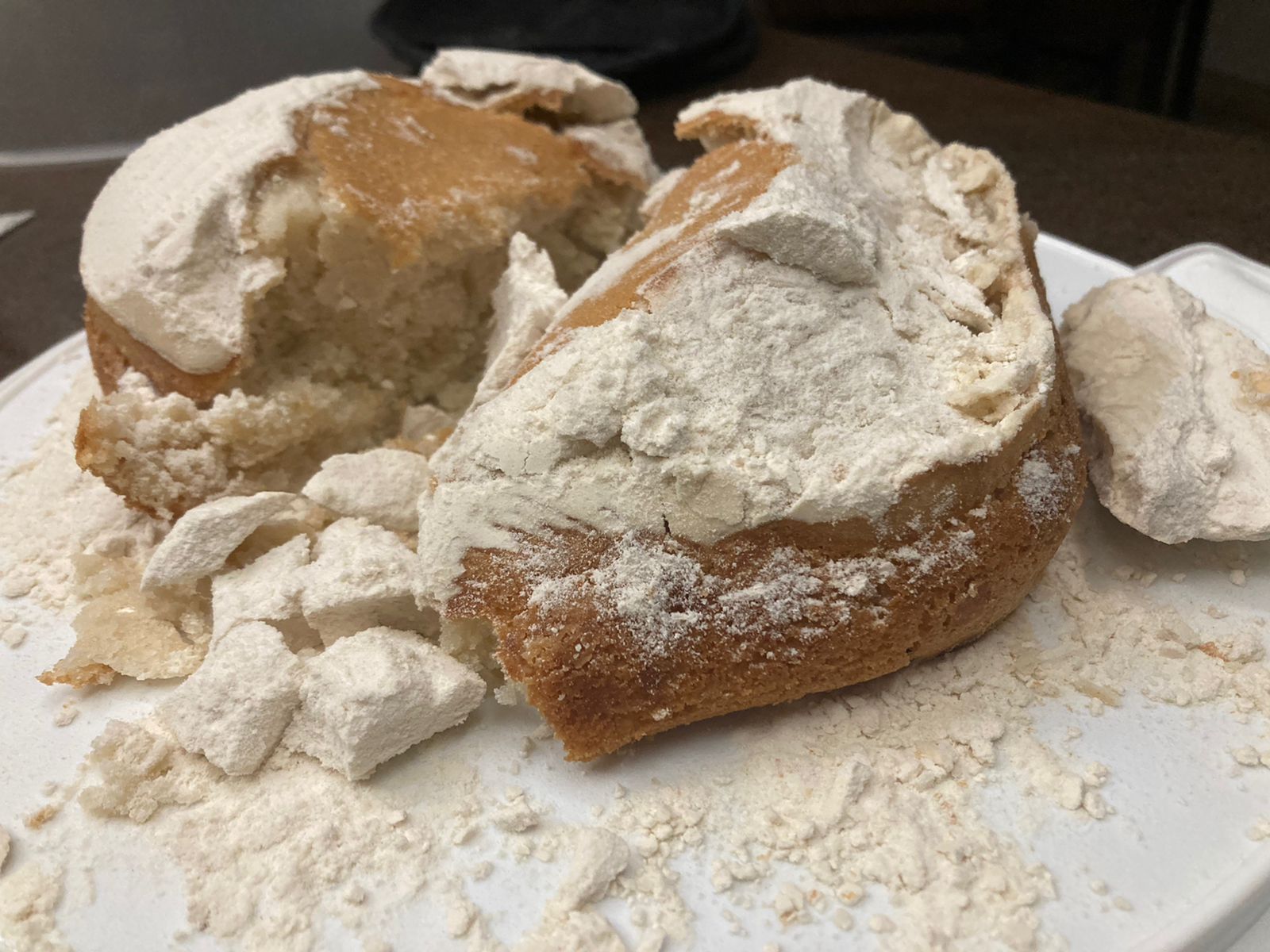 A round cake broken in half, covered with a thick layer of dry, white powder, with pieces and crumbs scattered around on a white plate.