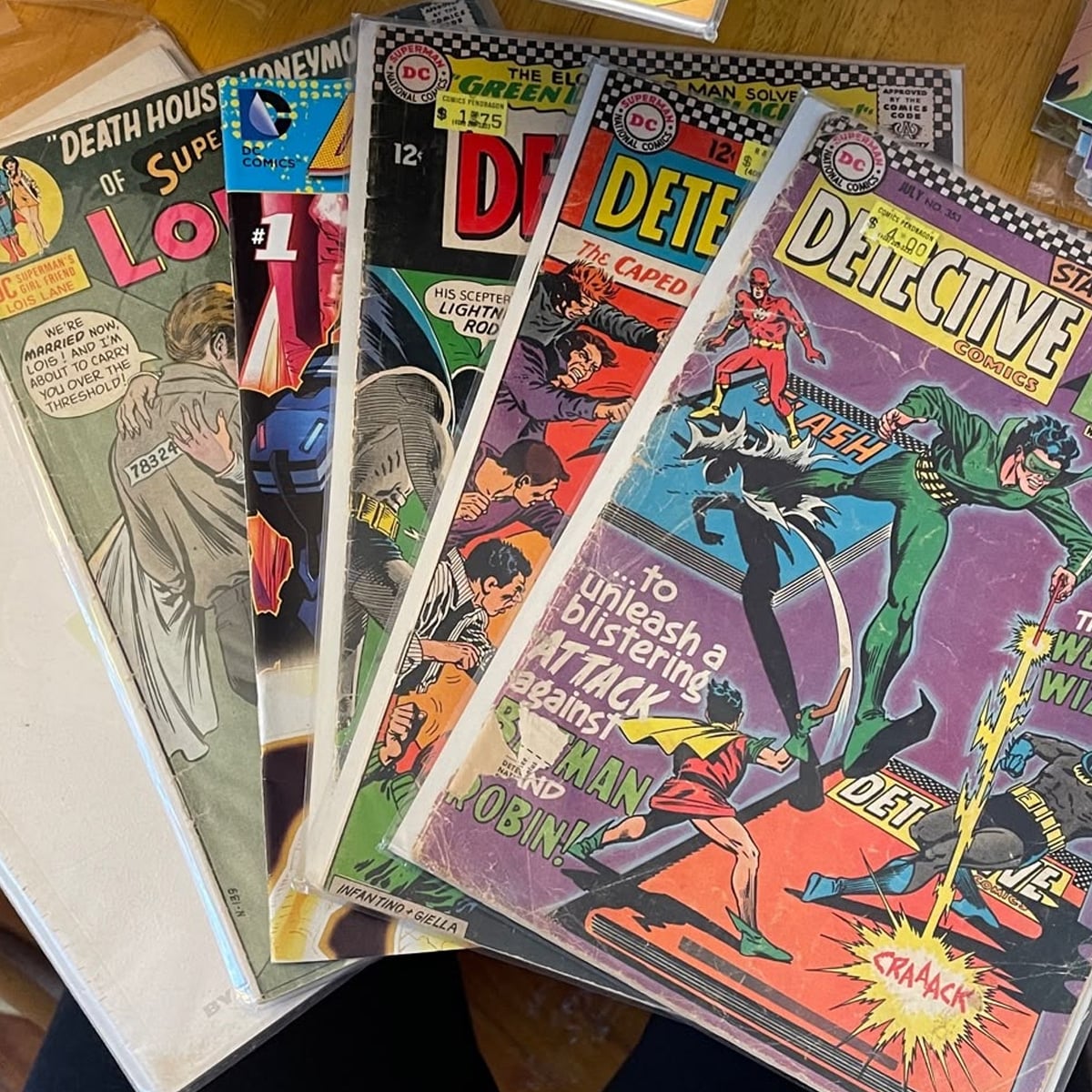 A fan spreads out several vintage comic books, including issues of Detective Comics and Superman’s Girlfriend Lois Lane, on a table. The colorful covers feature dramatic action scenes and classic superhero artwork.
