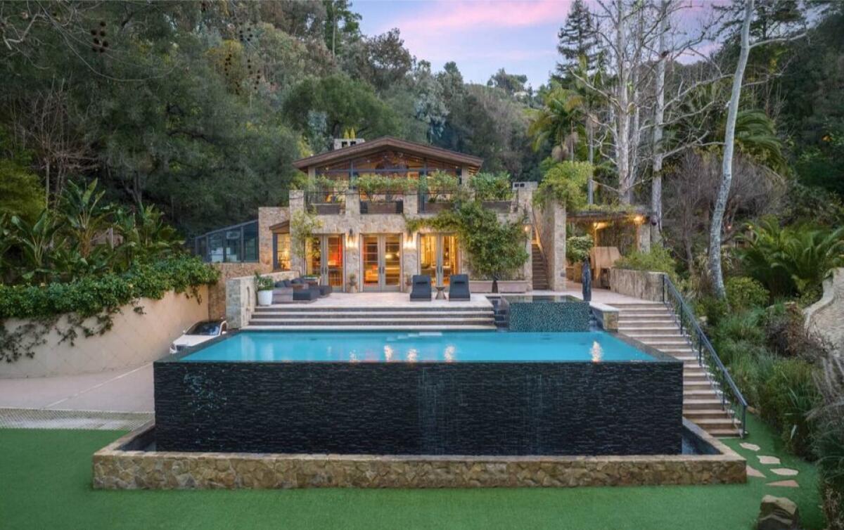 A modern two-story house with large windows and balconies is set amid lush trees. In front, there's a wide infinity pool with a stone border, outdoor seating, and steps leading to the house. The sky is tinged with pink at sunset.