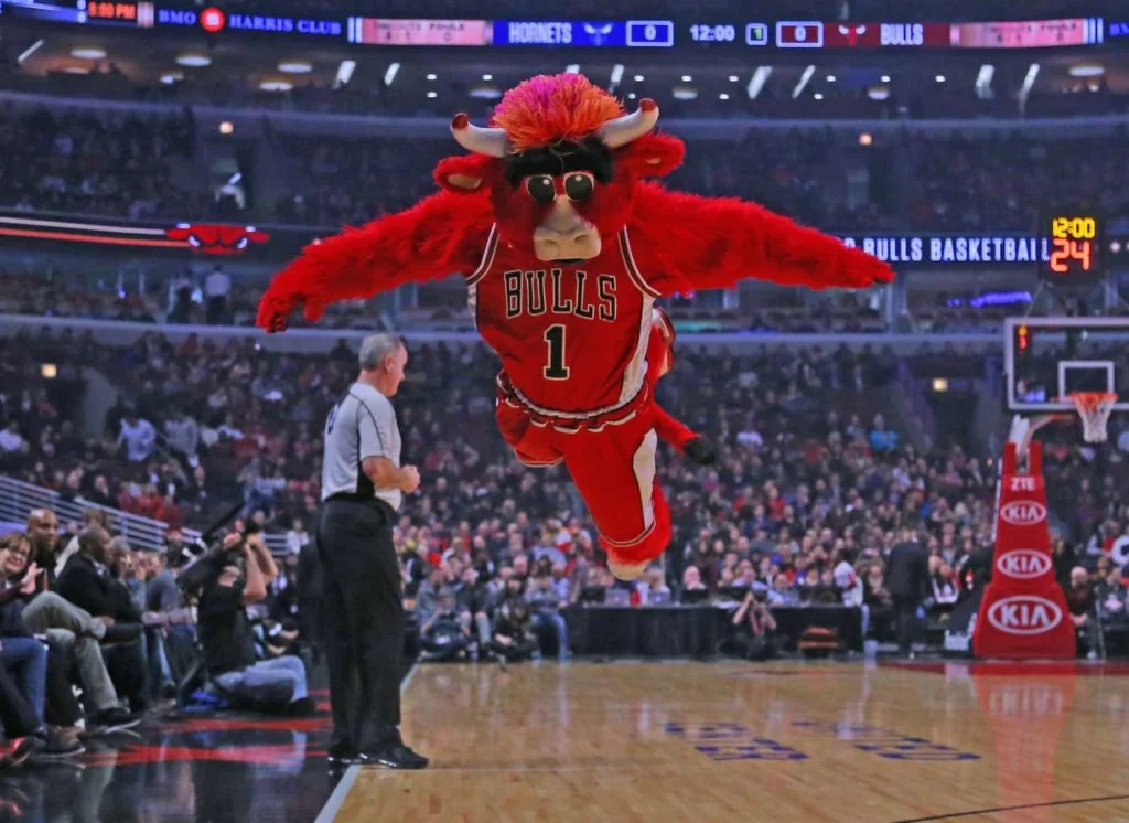 A person in a red bull mascot costume, wearing a "BULLS" jersey, is leaping through the air on a basketball court during a game, with fans and a referee watching in the background.